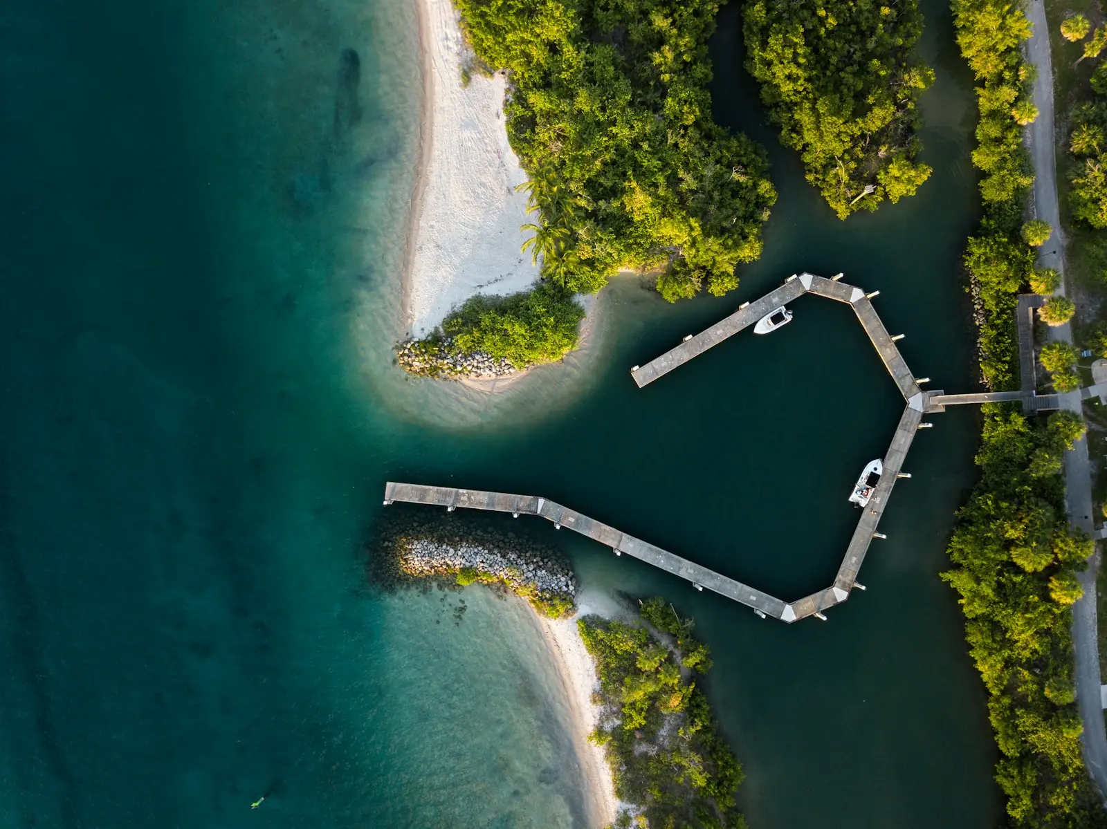 Aerial view of a curving dock, white sand and turquoise water beside a wooded shore